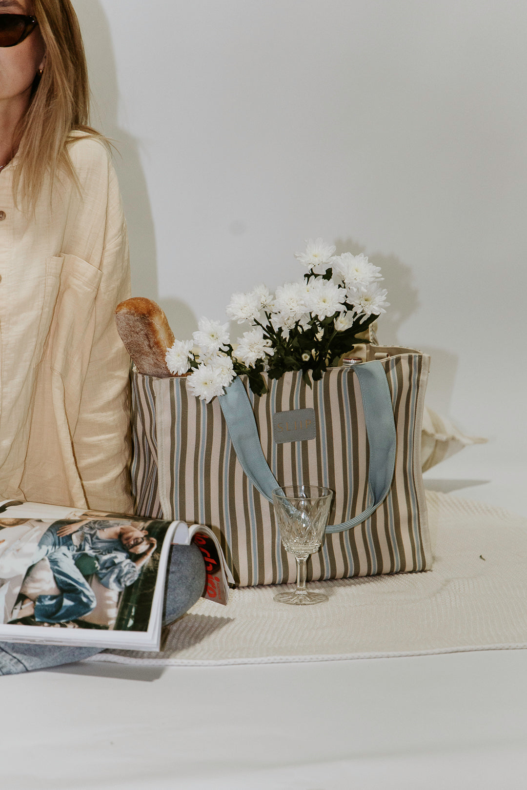 Women next to striped blue, green and beige tote bag with a light blue handles, bag filled with magazine, flowers and wine glass